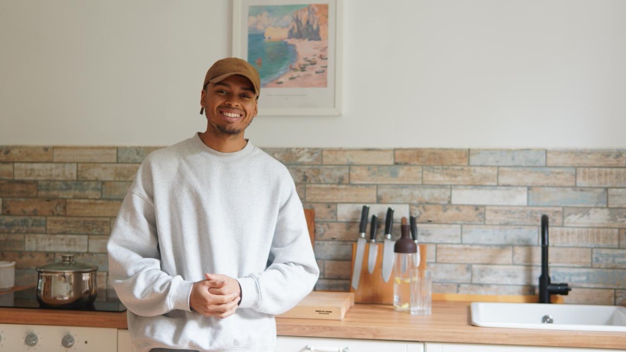 Eine lächelnde Person mit beiger Mütze und grauem Sweatshirt steht in einer modernen Küche mit hölzernen Arbeitsplatten, einem Messerblock und einem Topf auf dem Herd, hinter der ein Küstenkunstwerk an der Wand hängt.