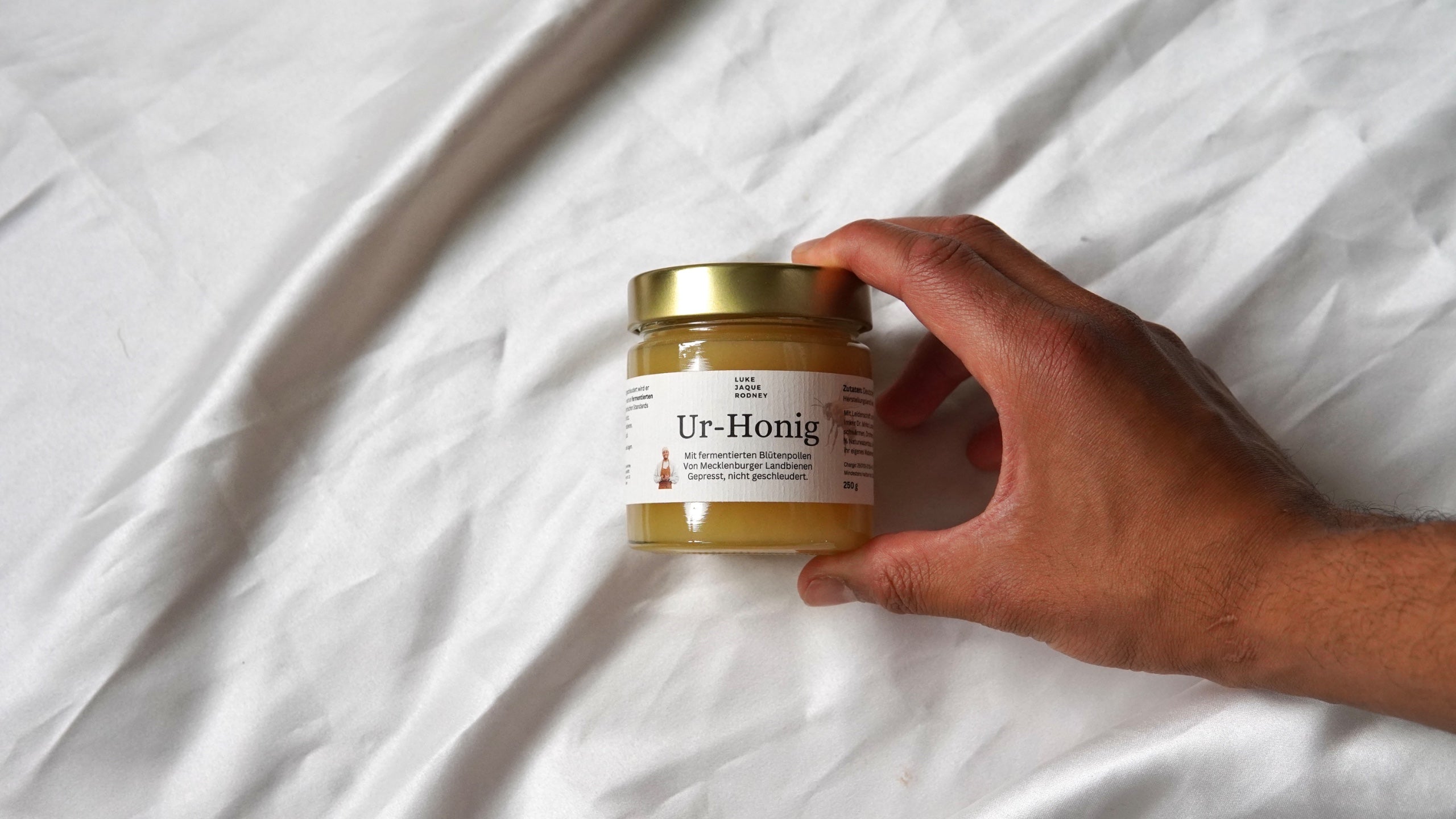 Eine Hand hält ein Glas Honig aus Ur-Honig mit goldenem Deckel und weißem Etikett vor einem weißen Stoffhintergrund.