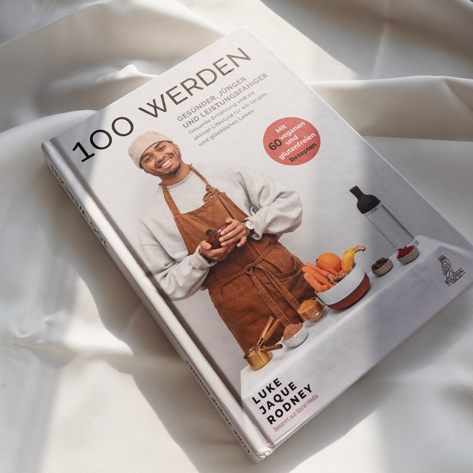 Das 100 werden Kochbuch | Bestseller von Luke Jaque Rodney Store ruht auf einem weißen Stoff. Sein Cover zeigt einen lächelnden Mann in einer braunen Schürze mit Gemüse, das gesunde Ernährung und Wohlbefinden symbolisiert, mit gesunden Lebensmitteln und Utensilien vor sich.