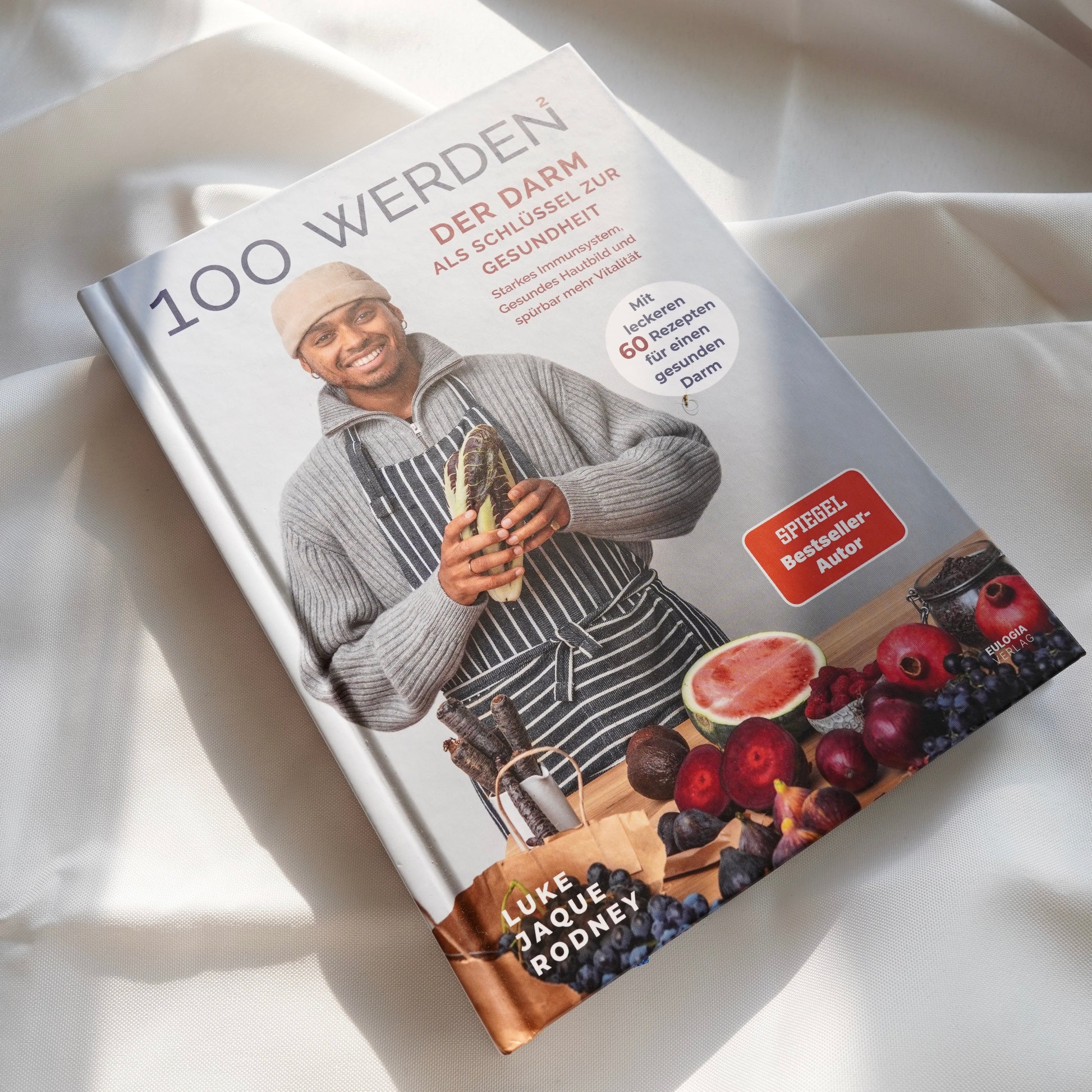 Das "100 werden 2 | Kochbuch" von Luke Jaque Rodney Store liegt auf leichtem Stoff. Sein Cover zeigt einen lächelnden Mann in Pullover und Schürze mit Gemüse, Feigen und Weintrauben, der auf gesunde Ernährung und Darmgesundheit für Langlebigkeit setzt.