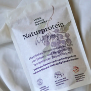 Ein weißer Beutel mit Naturprotein (Wild Berry) von Luke Jaque-Rodney mit Beeren-Illustrationen hebt veganes Protein aus Erbsen und Reis, fermentierten Beeren und Früchten der Saison hervor. Die Verpackung liegt auf einem weichen weißen Stoffhintergrund.