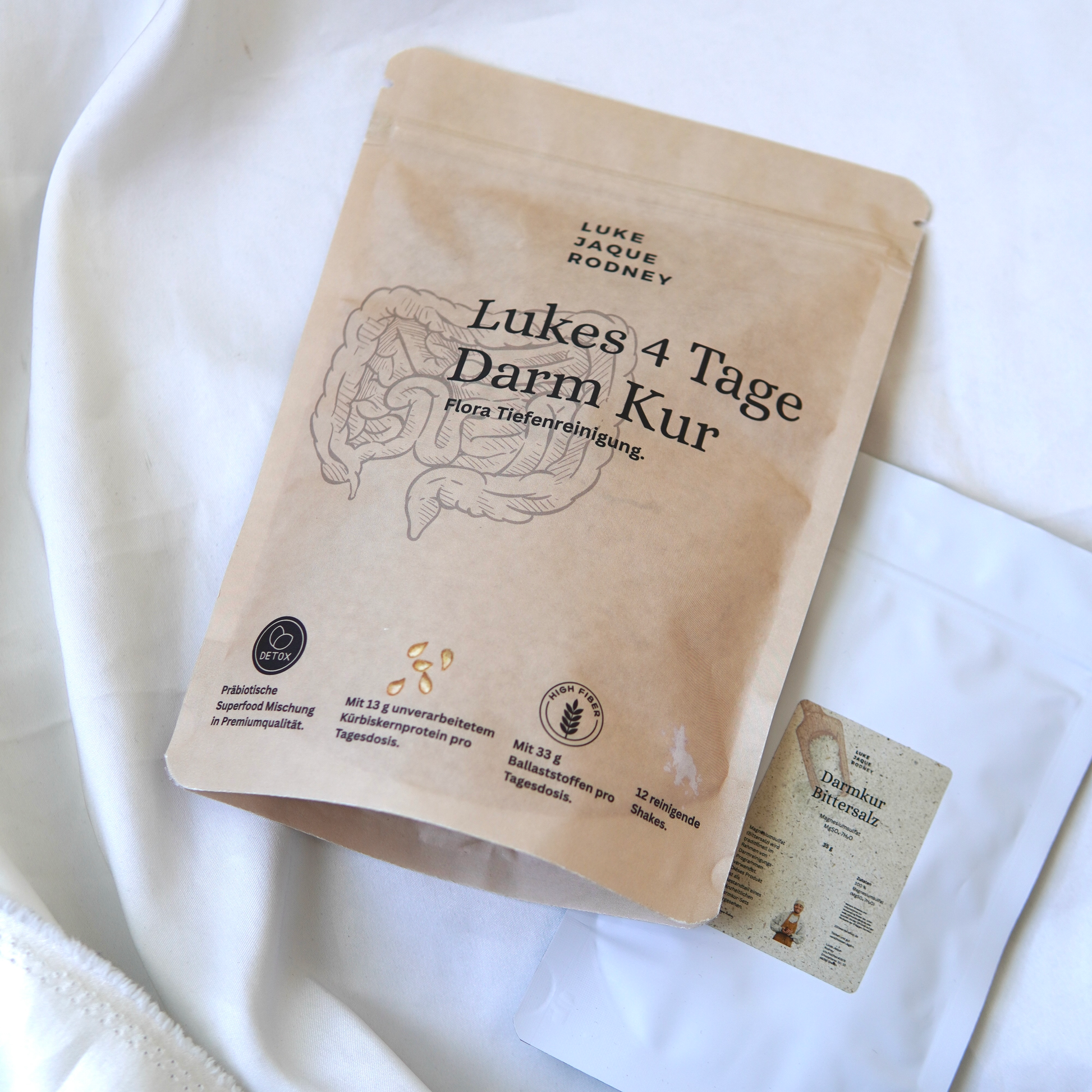 Ein braunes Päckchen mit der Aufschrift "Lukes 4-Tage-Darmkur Basic" von Luke Jaque-Rodney, auf dem Darmbilder abgebildet sind, liegt neben einem kleineren weißen Tütchen. Der deutsche Text hebt Ballaststoffe, Probiotika und Darmreinigung zur Reinigung der Darmflora hervor.