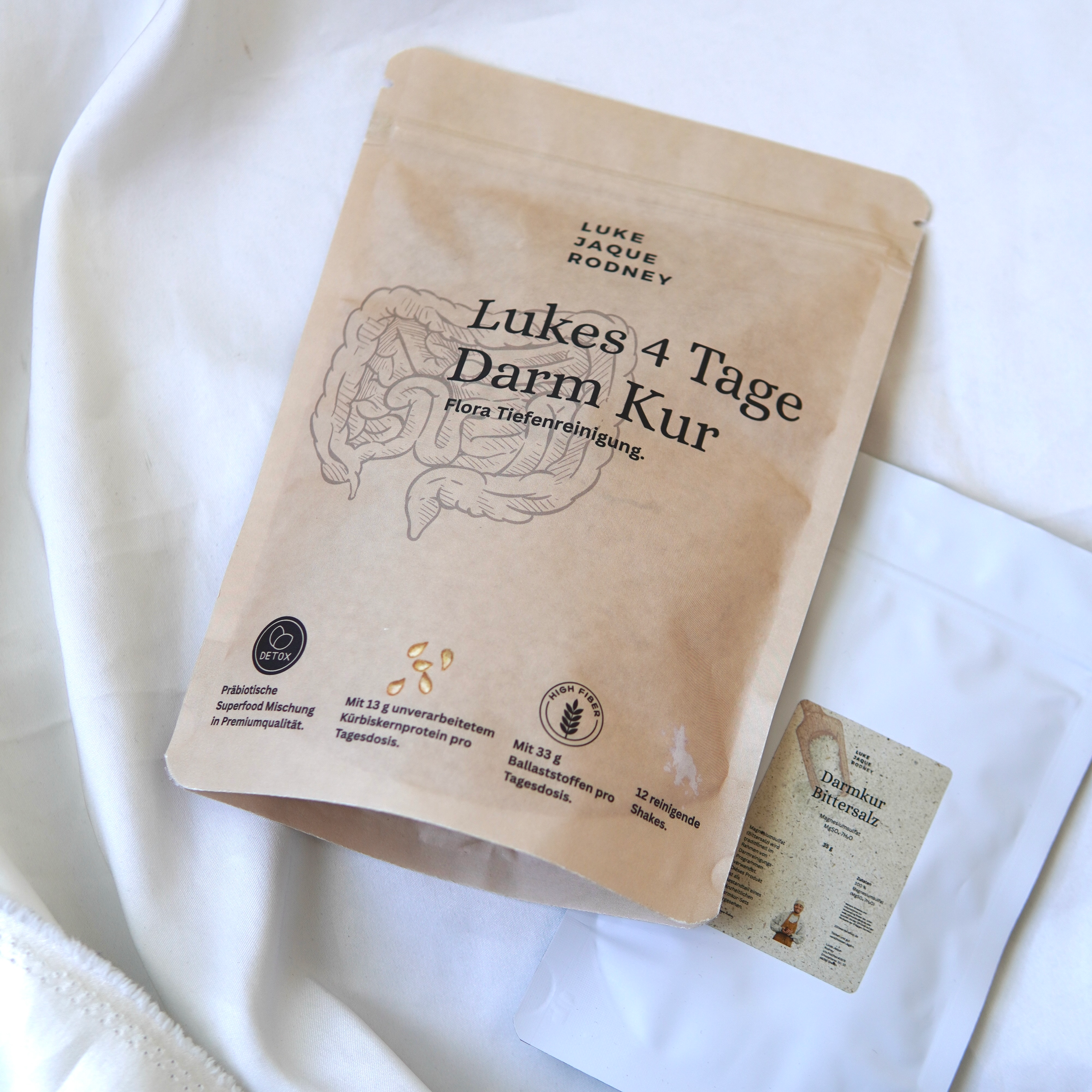 Ein braunes Päckchen mit der Aufschrift "Lukes 4-Tage-Darmkur Basic" von Luke Jaque-Rodney, auf dem Darmbilder abgebildet sind, liegt neben einem kleineren weißen Tütchen. Der deutsche Text hebt Ballaststoffe, Probiotika und Darmreinigung zur Reinigung der Darmflora hervor.