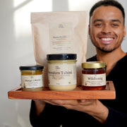 Ein lächelnder Mann hält ein Holztablett mit Gläsern und einer Tüte in der Hand, in der das Lukes Starter Bundle von Luke Jaque-Rodney präsentiert wird - eine Auswahl an köstlichen Bio-Lebensmitteln wie Omega-3-Butter, Premium-Tahini, Wildhonig, Sukkari-Datteln und Naturbonbons.