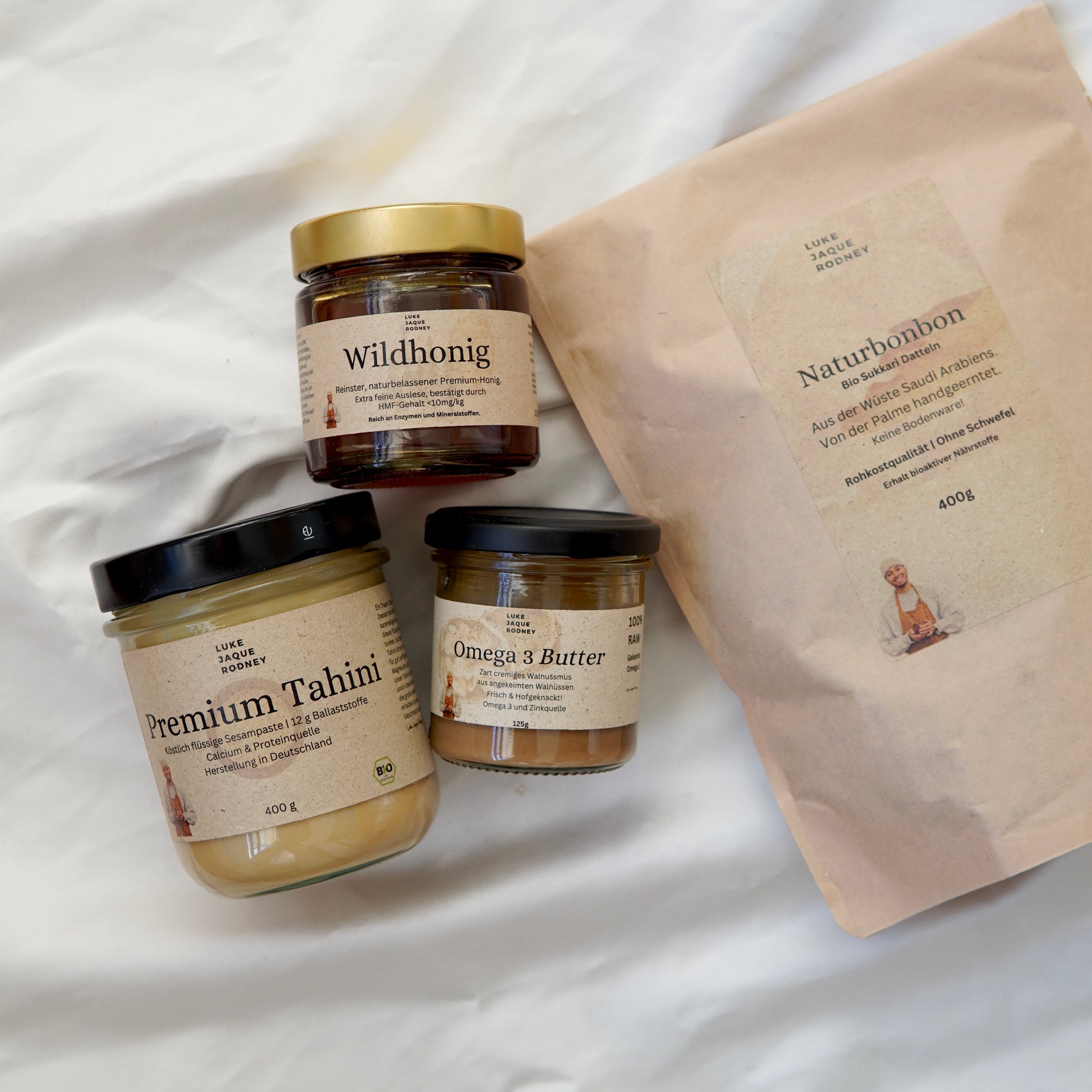 Drei Gläser aus Luke Jaque-Rodneys Lukes Starter Bundle sind neben einer braunen Naturbonbon-Papiertüte auf weißem Stoff arrangiert - ideal zum Verschenken oder zum Kombinieren mit Sukkari-Datteln.