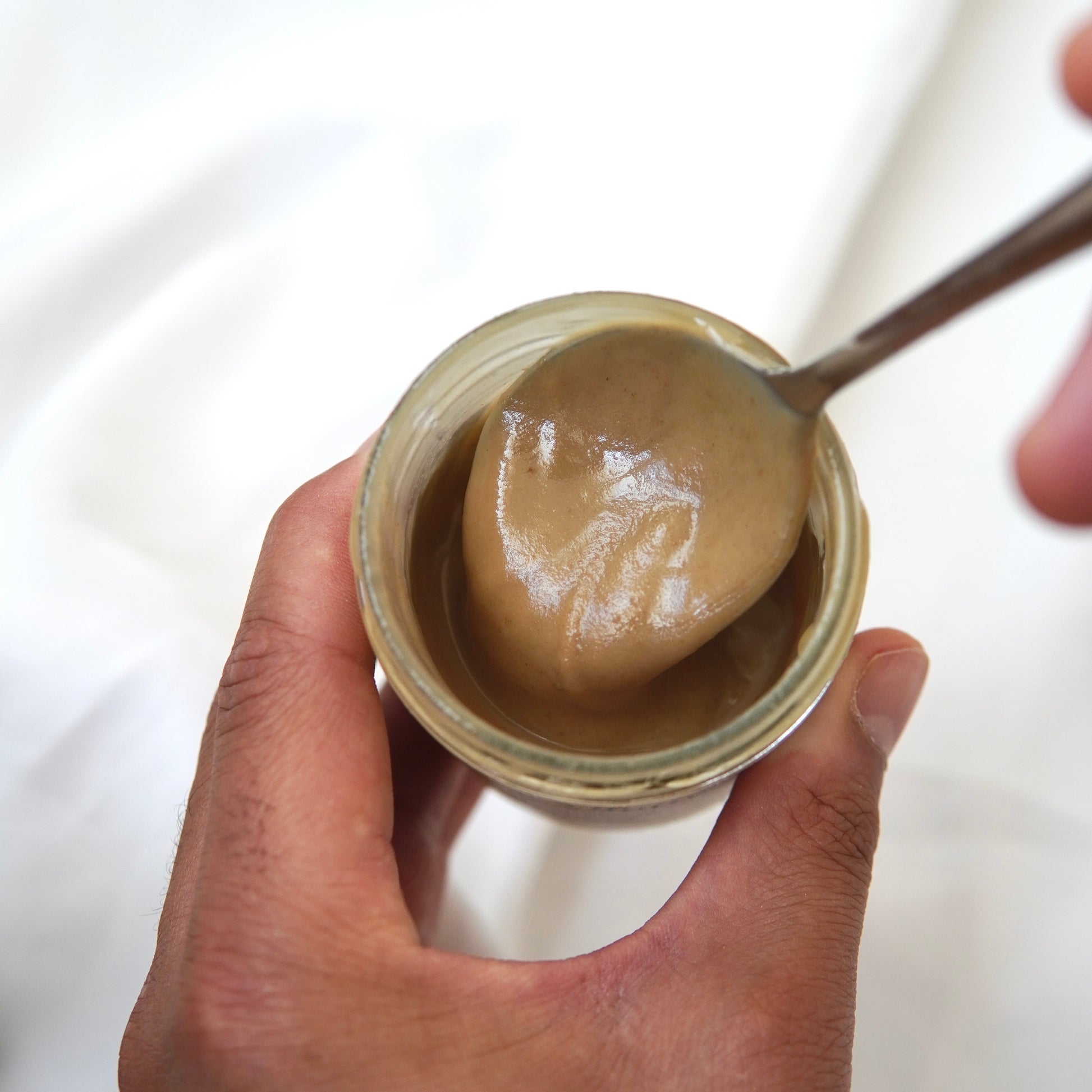 Eine Hand hält ein Glas Omega-3-Butter I Walnussmus von Luke Jaque-Rodney, während eine andere Hand mit einem Löffel etwas davon vor einem weißen Hintergrund schöpft.