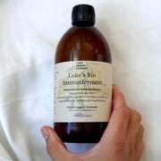 Eine Hand hält eine braune Glasflasche mit der Aufschrift Lukes Bio Immunferment I Rohkost Essenz aus dem Luke Jaque-Rodney Store, die ein weißes und beiges deutsches Etikett mit der Aufschrift Bio Zitronen Ingwer Kurkuma Saft und fermentierte Essenz vor einem weißen Hintergrund aufweist.
