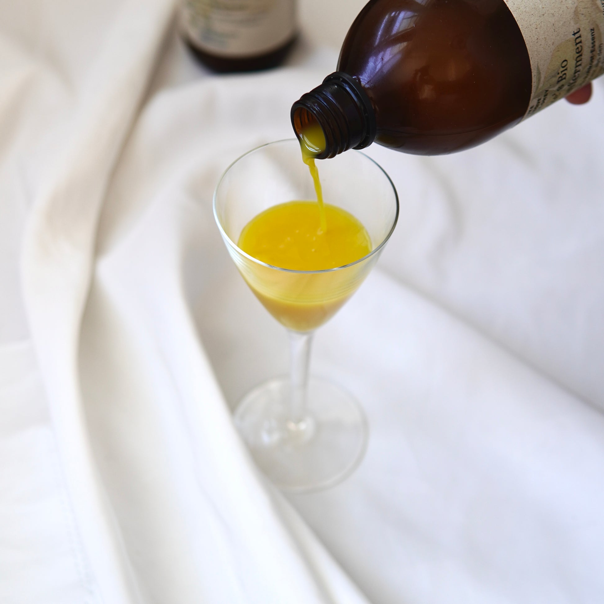 Eine braune Flasche gießt eine gelbe Flüssigkeit, Lukes Bio Immunferment I Rohkost Essenz von Luke Jaque-Rodney Store, in ein klares Stielglas auf einem weißen Stoffhintergrund.