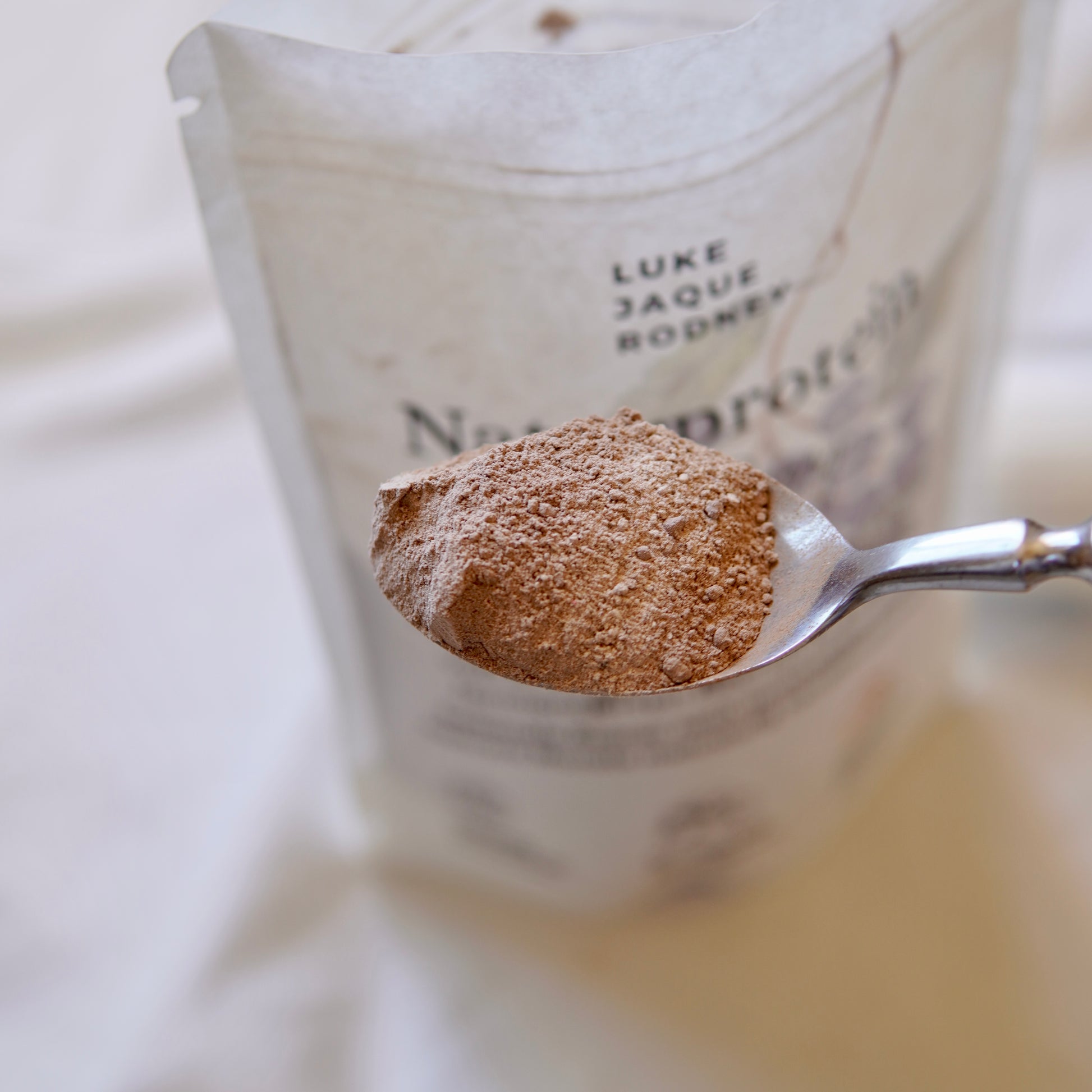 Eine Nahaufnahme zeigt einen Metalllöffel mit einem Haufen des veganen Proteinpulvers Naturprotein (Wild Berry) von Luke Jaque-Rodney vor einem verschwommenen Proteinpulverbeutel auf einer hellen Fläche.