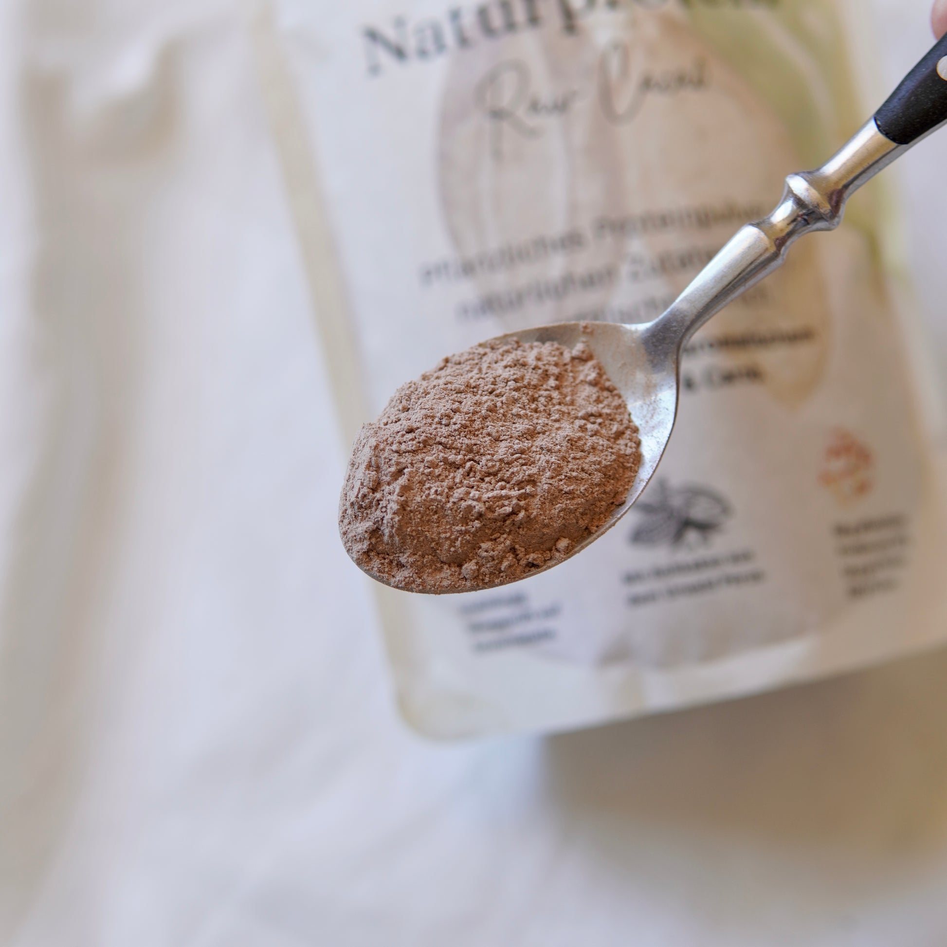 Eine Nahaufnahme eines Löffels mit hellbraunem Naturprotein (Raw Cacao) von Luke Jaque-Rodney, mit einer unscharfen Verpackung des veganen Proteins im Hintergrund auf einer weißen Fläche.