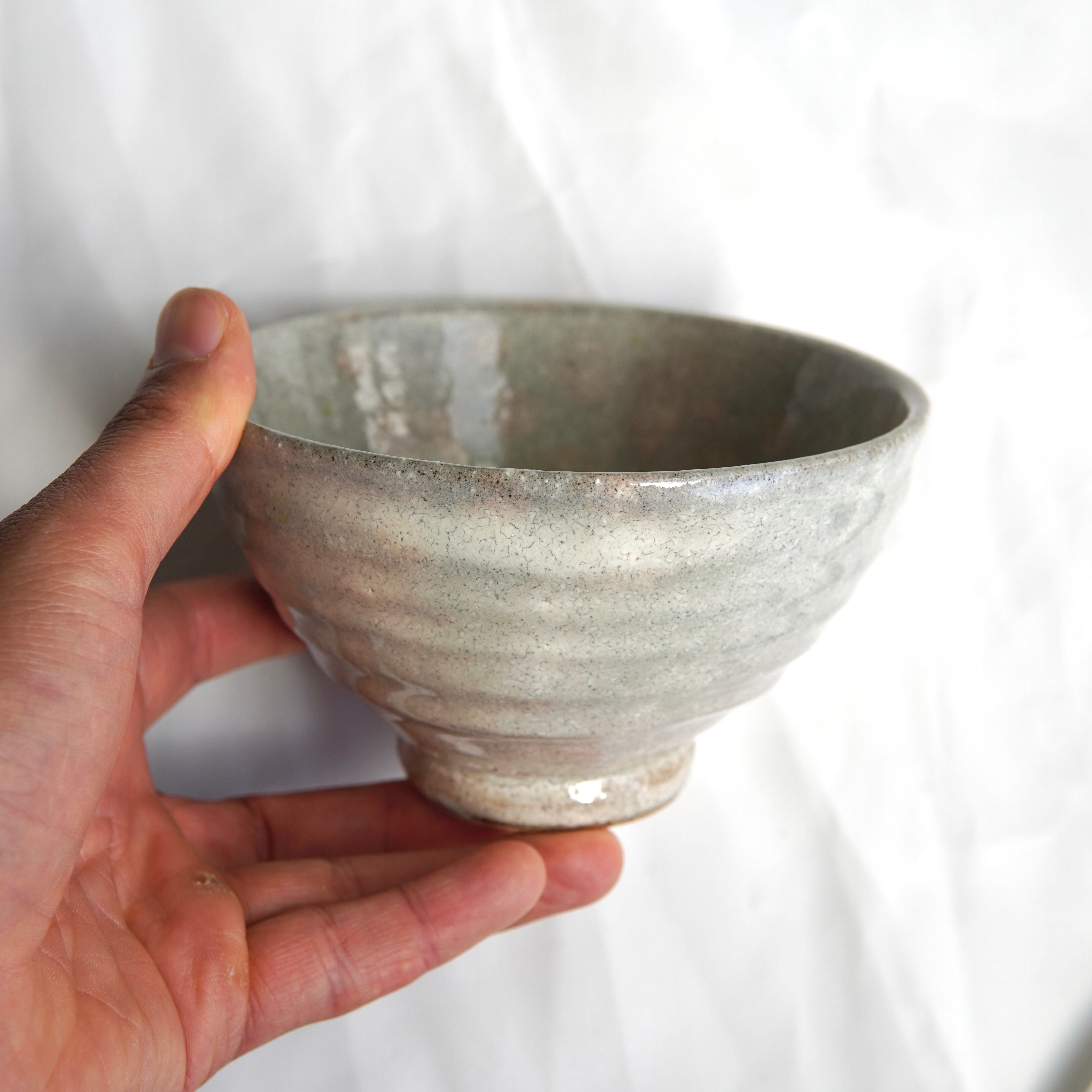 Eine Hand hält eine strukturierte, hellgraue Matcha-Schale aus dem Matcha-Set | Handgefertigt von Luke Jaque Rodney Store, mit leicht unebener Oberfläche und kleinem Fuß, vor weißem Hintergrund.