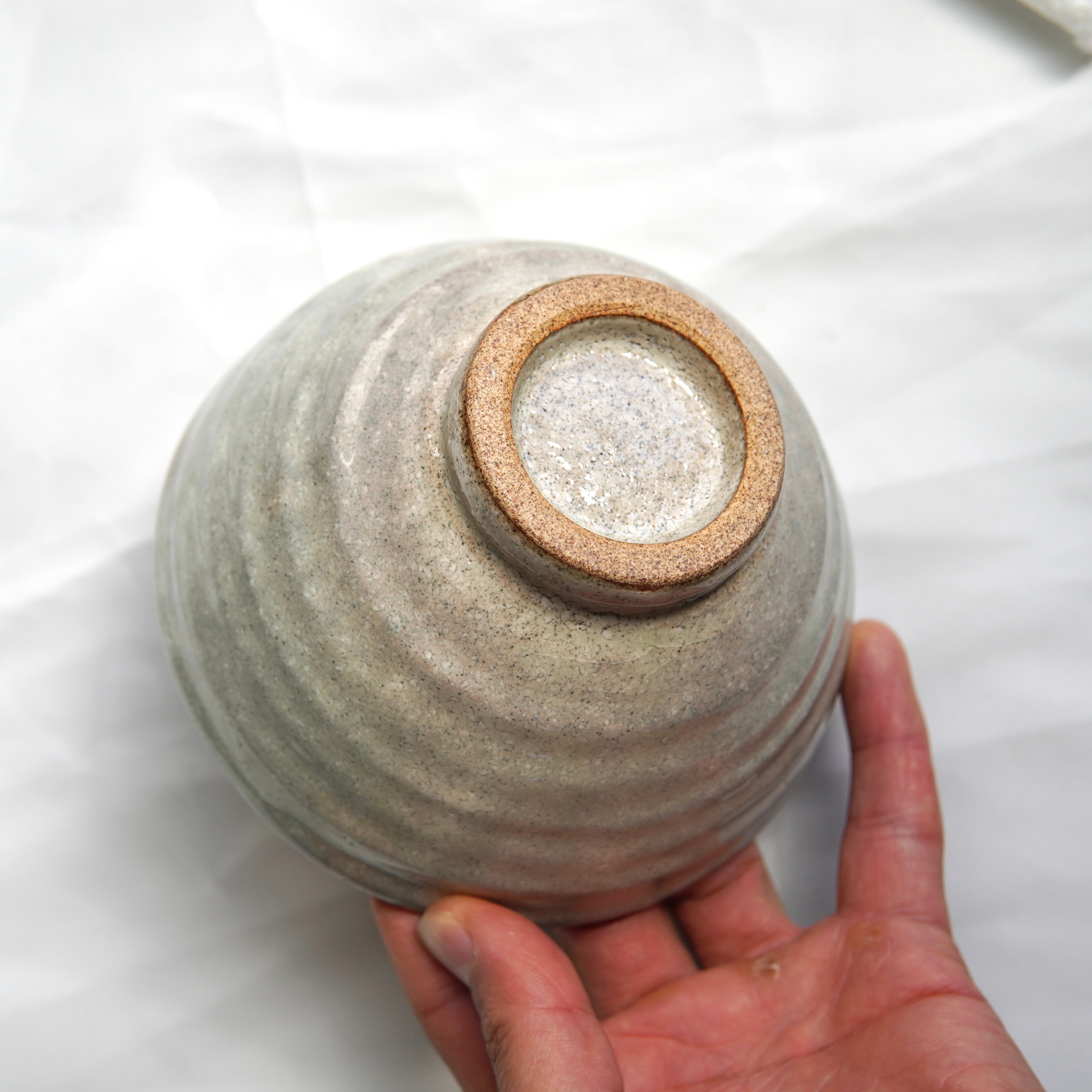 Eine Hand hält eine umgedrehte Matcha-Schale aus dem Matcha-Set | Handgefertigt von Luke Jaque Rodney Store, mit einer strukturierten hellgrauen Oberfläche und einem unglasierten runden Sockel vor weißem Hintergrund.