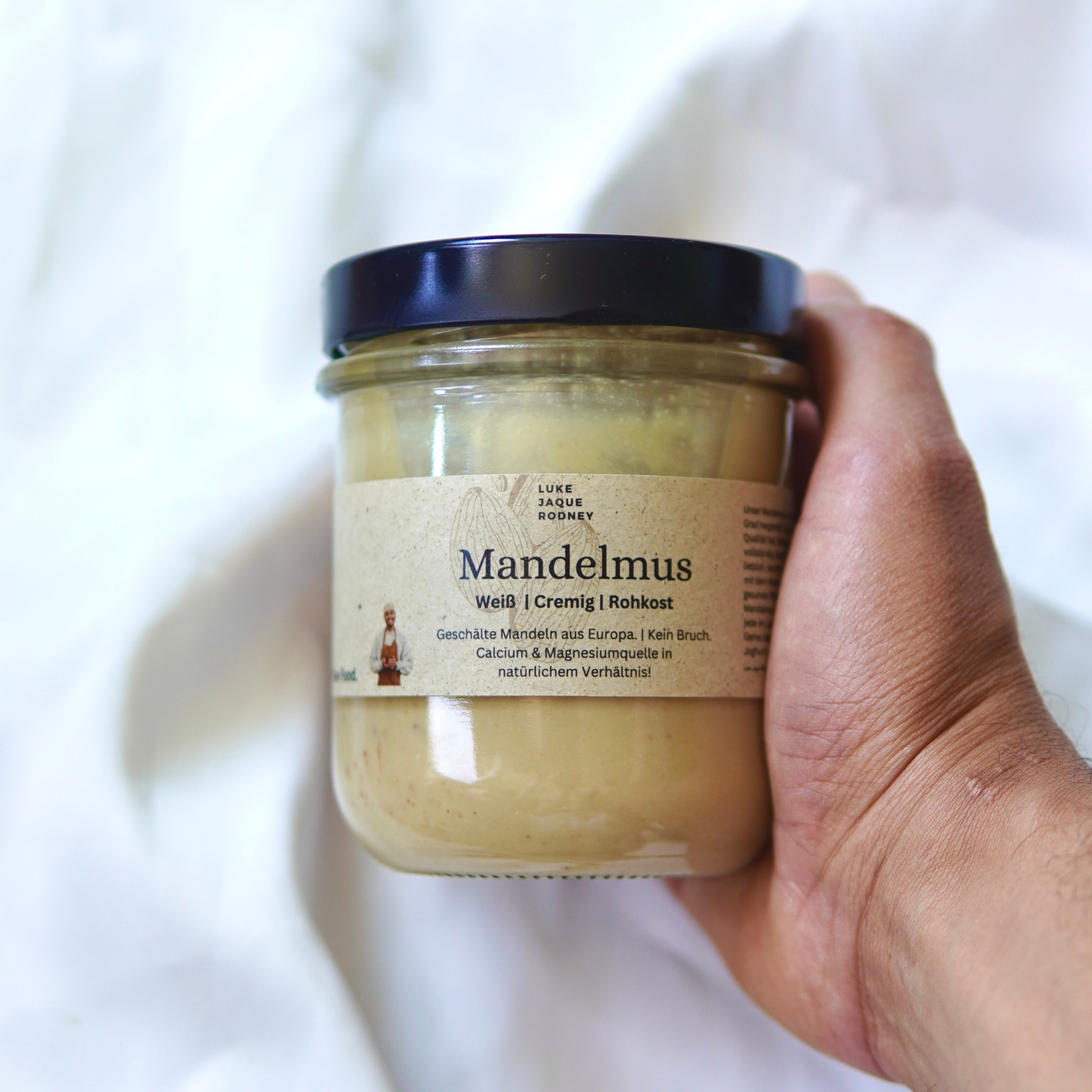 Eine Hand hält ein Glas mit Luke Jaque Rodney Store Mandelmus I Weiß, Rohkost mit einem dunklen Deckel. Auf dem deutschen Etikett steht, dass es roh und aus europäischen Mandeln hergestellt ist - ideal für Pflanzenmilch. Ein sanft verschwommener Hintergrund lenkt die Aufmerksamkeit auf das Glas.