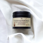 Eine schwarze Dose mit der Aufschrift Matcha | Ceremonial Grade | Kagoshima aus dem Luke Jaque Rodney Store ruht auf einem weiß drapierten Stoff. Das beigefarbene, deutschsprachige Etikett hebt die antioxidative Kapazität hervor und zeigt einen Matcha-Besen.