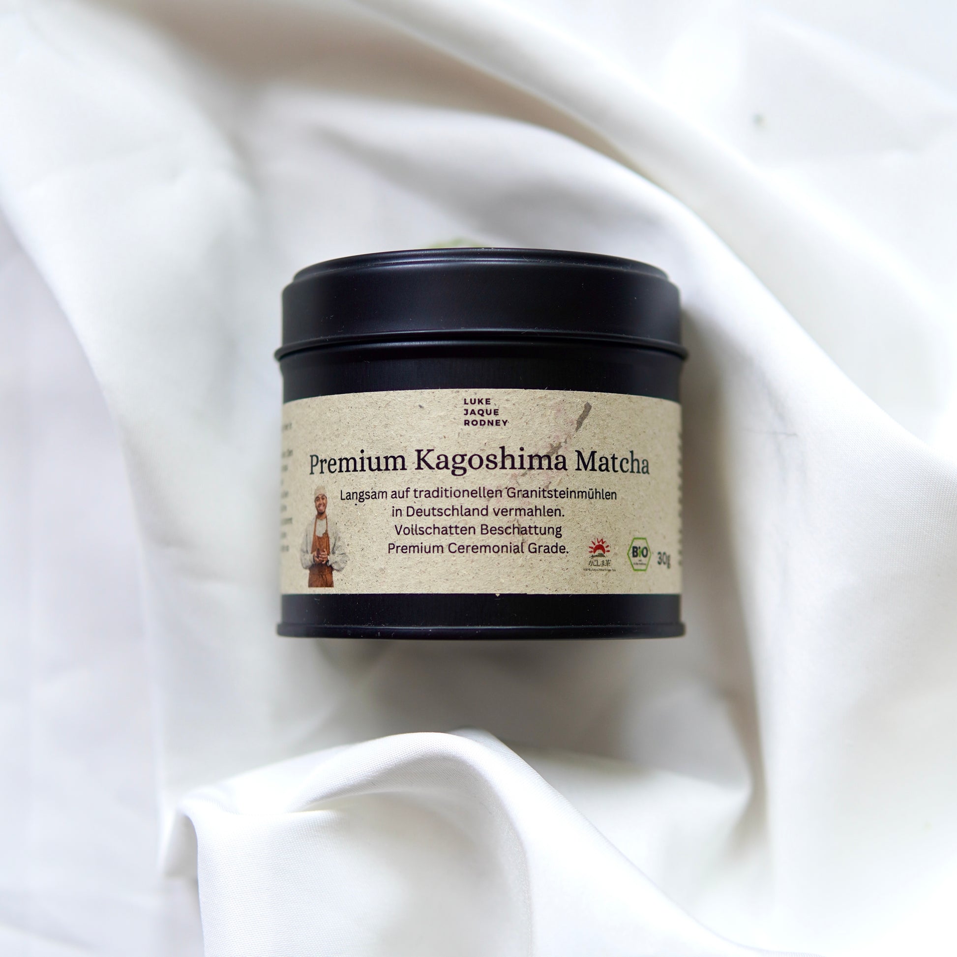 Eine schwarze Dose mit der Aufschrift Matcha | Ceremonial Grade | Kagoshima aus dem Luke Jaque Rodney Store ruht auf einem weiß drapierten Stoff. Das beigefarbene, deutschsprachige Etikett hebt die antioxidative Kapazität hervor und zeigt einen Matcha-Besen.