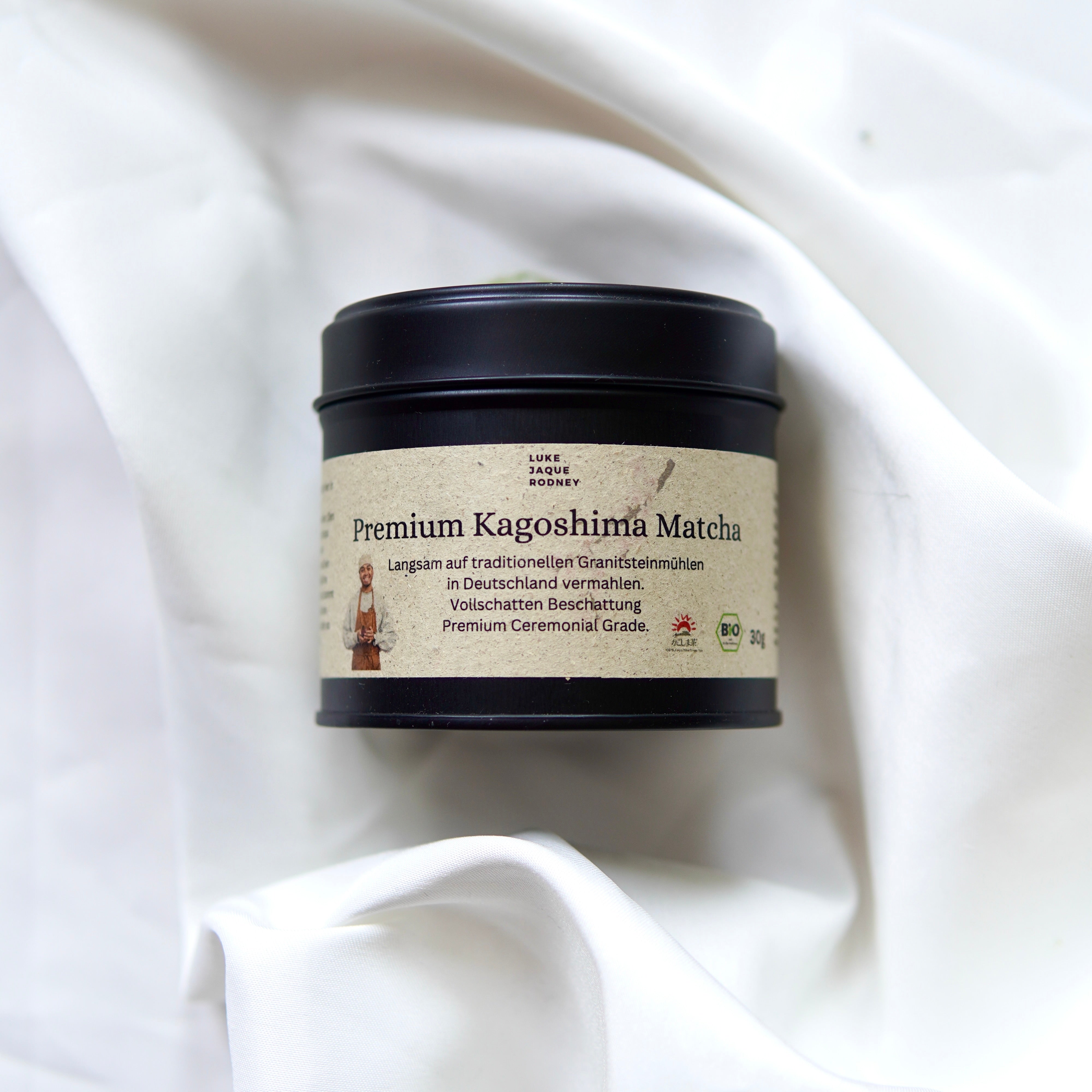 Eine schwarze Dose mit der Aufschrift Matcha | Ceremonial Grade | Kagoshima aus dem Luke Jaque Rodney Store ruht auf einem weiß drapierten Stoff. Das beigefarbene, deutschsprachige Etikett hebt die antioxidative Kapazität hervor und zeigt einen Matcha-Besen.