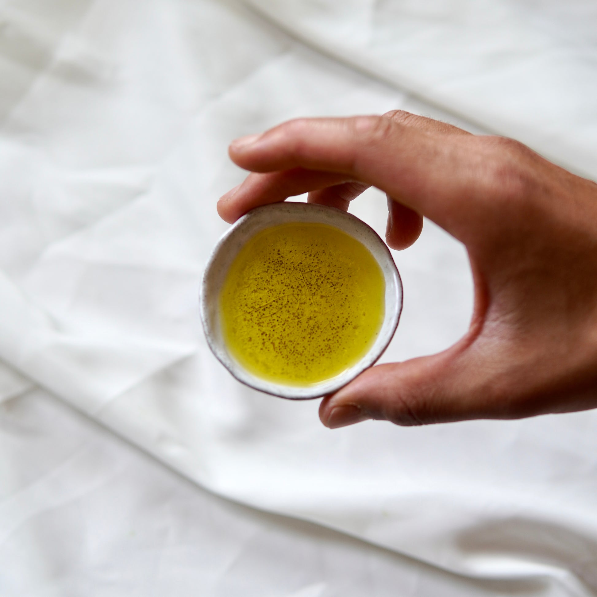 Eine Hand hält eine Flasche Das heilige Öl, ein Premium-Olivenöl aus dem Luke Jaque Rodney Store, das seinen hohen Polyphenolgehalt und seinen feinen goldenen Farbton vor einem weichen weißen Stoffhintergrund präsentiert.
