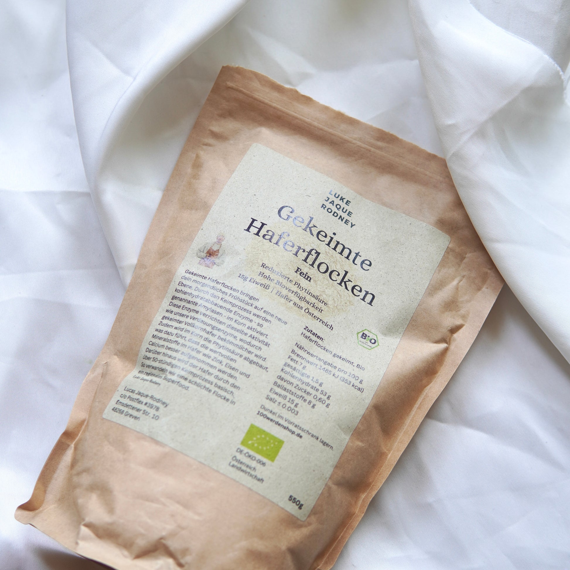 Eine braune Papierverpackung von Gekeimte Haferflocken | Fein und Grob von Luke Jaque Rodney Store, ein glutenfreies Superfood, ruht auf weißem Stoff. Das Etikett zeigt die Bio-Zertifizierung und deutsche Produktinformationen.