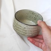 Eine Hand hält eine runde, strukturierte Matcha-Schale aus dem Matcha-Set | Handgefertigt von Luke Jaque Rodney Store mit einer hellen grünlich-grauen Glasur vor einem weichen weißen Stoffhintergrund.