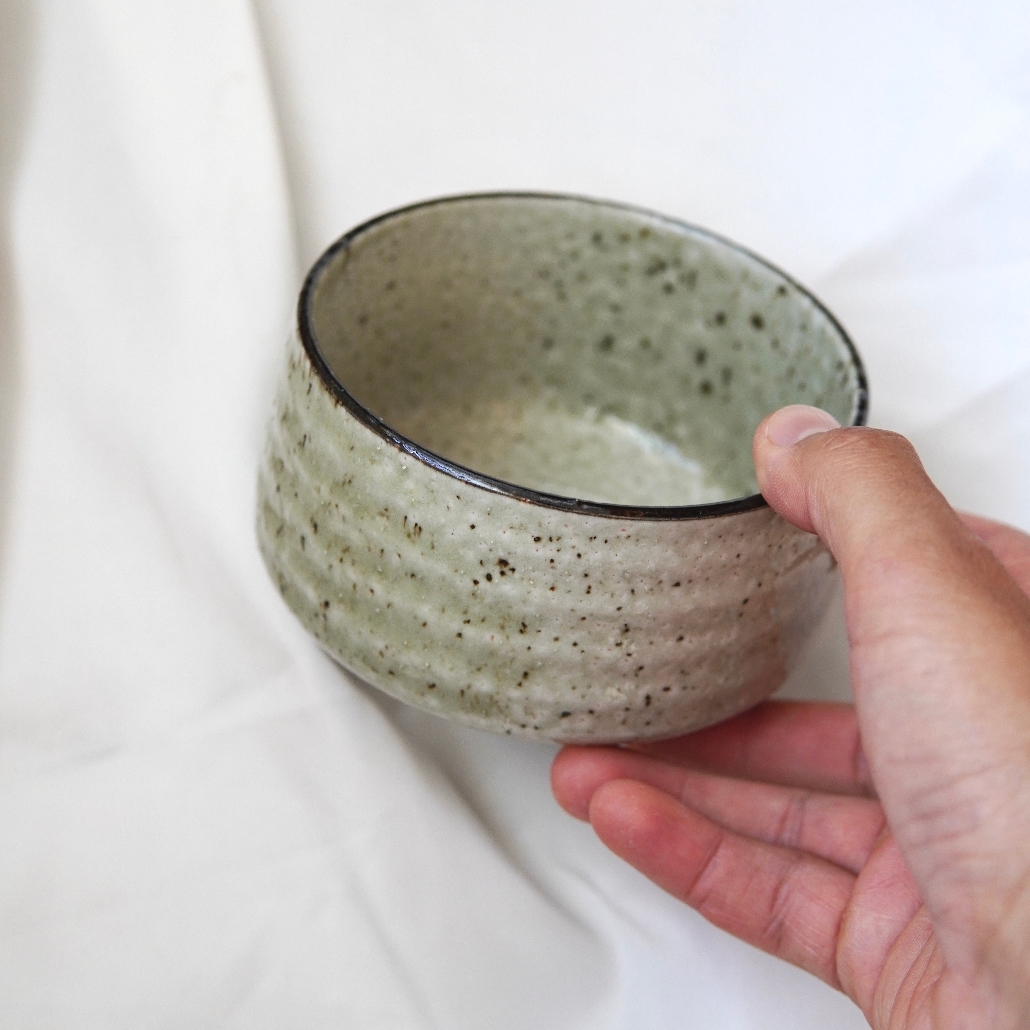 Eine Hand hält eine runde, strukturierte Matcha-Schale aus dem Matcha-Set | Handgefertigt von Luke Jaque Rodney Store mit einer hellen grünlich-grauen Glasur vor einem weichen weißen Stoffhintergrund.