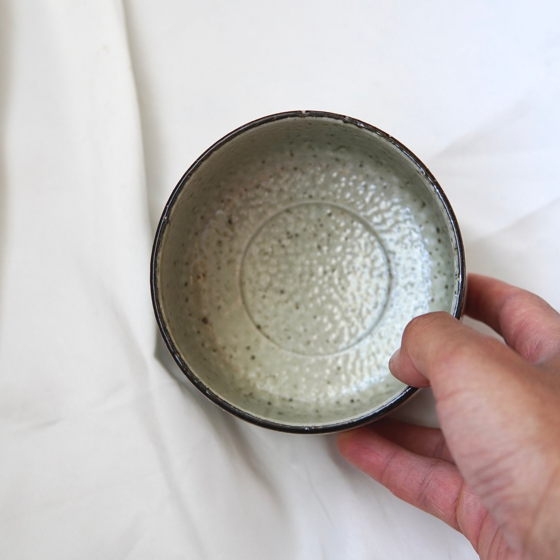 Eine Hand hält das Matcha-Set | Handgefertigt von Luke Jaque Rodney Store - ein runder, gesprenkelter Chawan aus Keramik mit einem strukturierten Inneren, von oben gesehen vor einem weißen Stoffhintergrund.