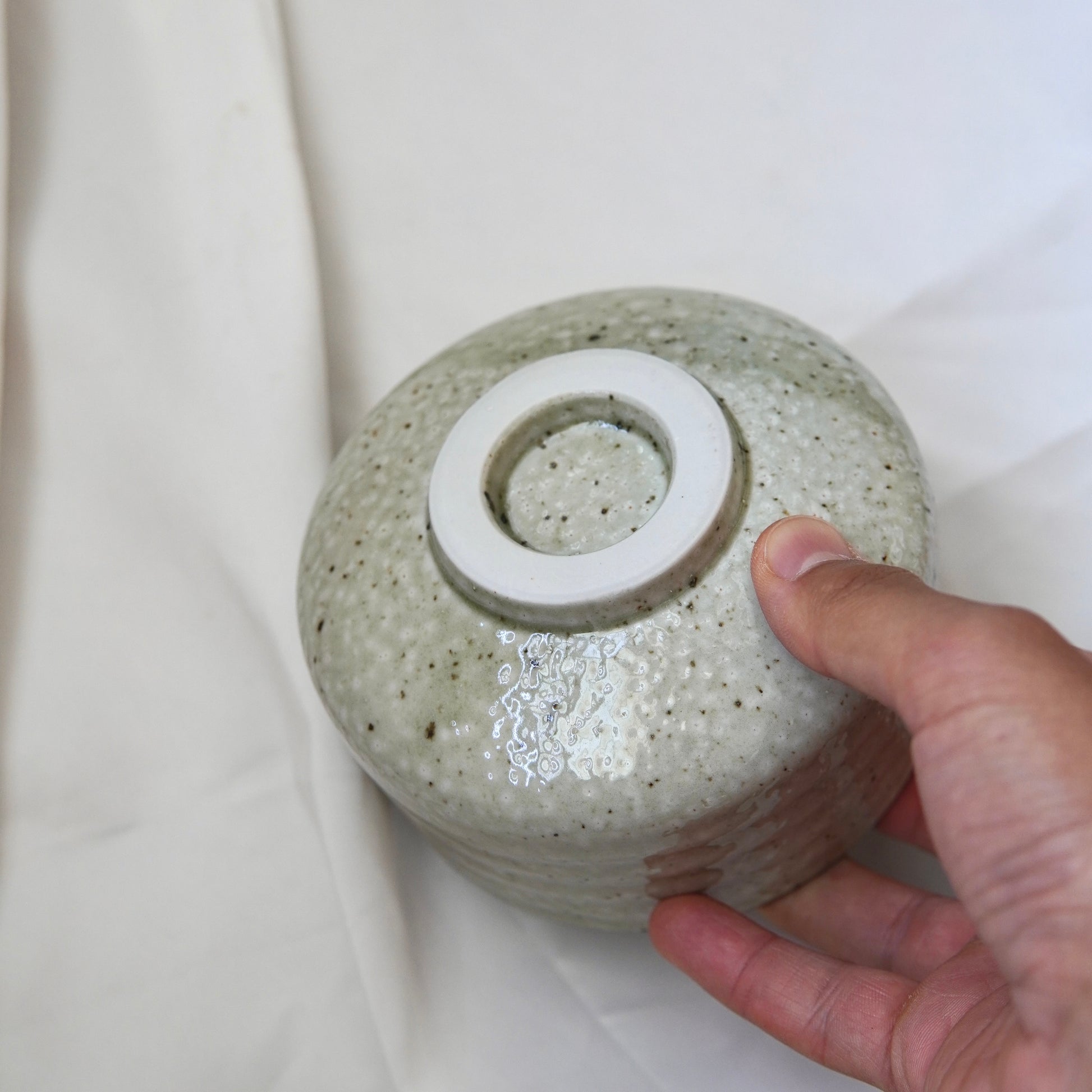 Eine Hand hält eine umgedrehte, hell gesprenkelte Keramikschale aus dem Matcha-Set | Handgefertigt von Luke Jaque Rodney Store, deren kreisrunder Fuß auf einem weißen Stoffhintergrund zu sehen ist.