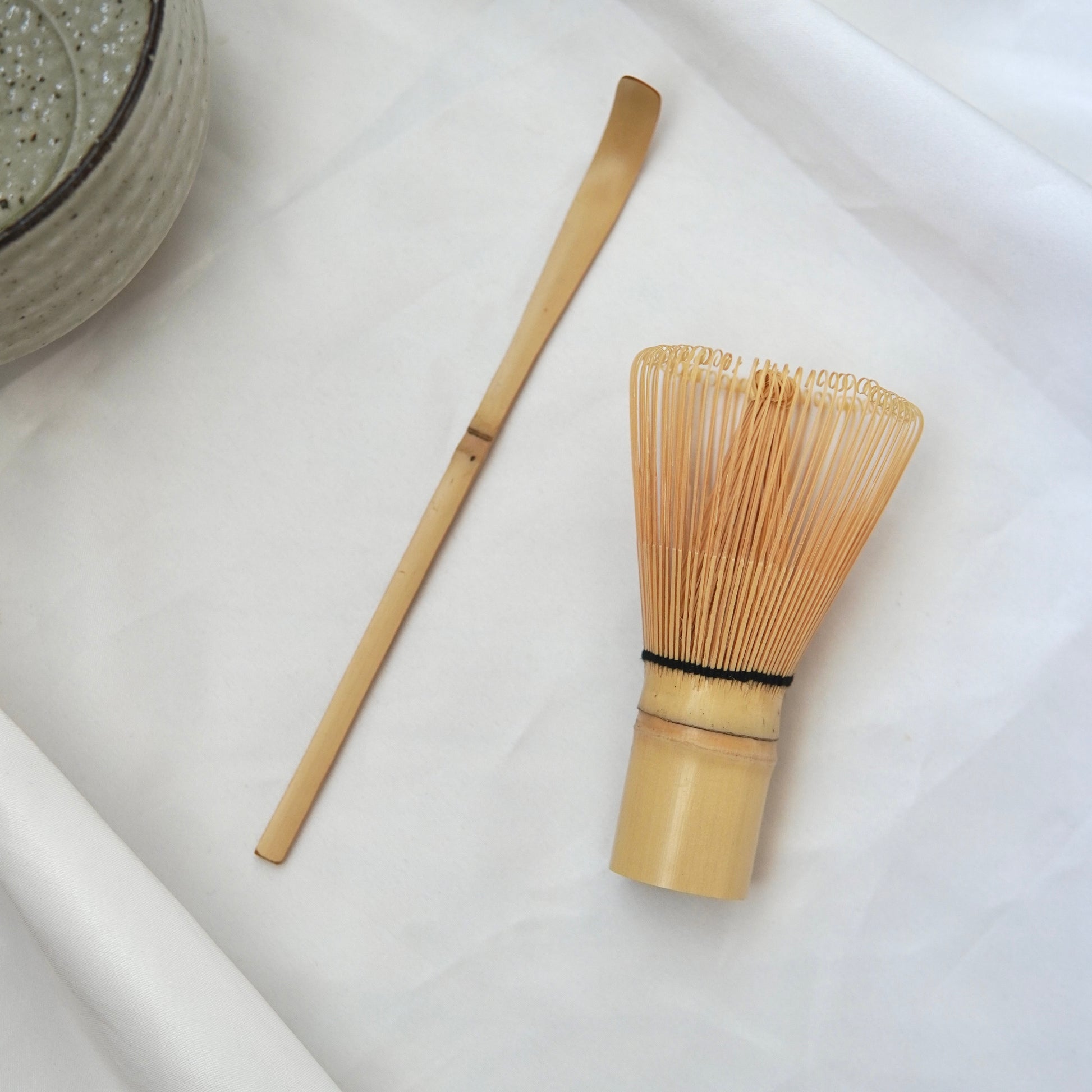 Das handgefertigte Matcha-Set von Luke Jaque Rodney Store mit Bambusbesen und -löffel liegt auf weißem Tuch neben einer Matcha-Schale - perfekt für die Zubereitung deines Matchas.