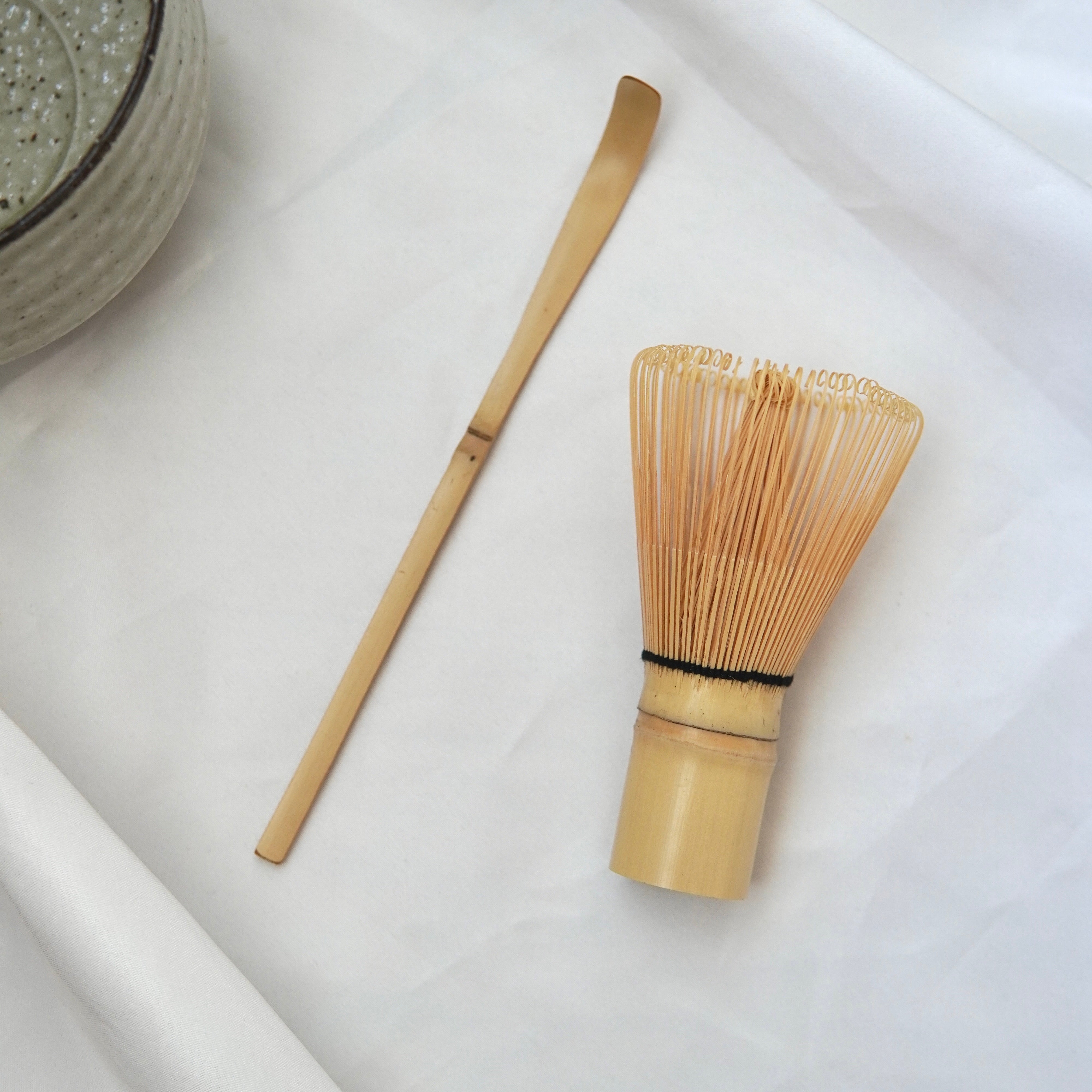 Das handgefertigte Matcha-Set von Luke Jaque Rodney Store mit Bambusbesen und -löffel liegt auf weißem Tuch neben einer Matcha-Schale - perfekt für die Zubereitung deines Matchas.