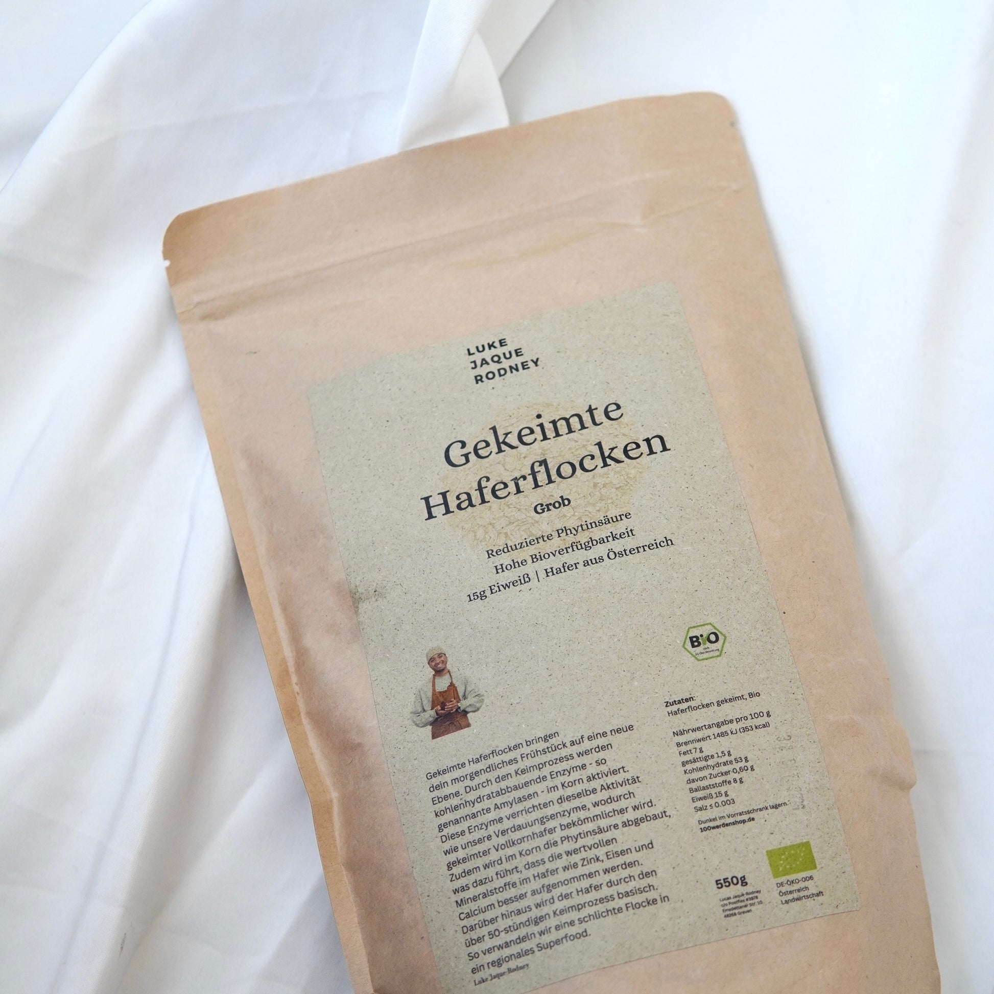 Eine braune Papierverpackung mit Gekeimte Haferflocken von Luke Jaque Rodney Store, beschriftet in deutscher Sprache mit Produktdetails, Logo, Zertifizierungssymbol und dem Bild einer Person unten links, liegt auf weißem Stoff.