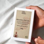 Eine Hand hält eine weiße Schachtel von Datteln | Mazafati Permakultur von Luke Jaque Rodney Store, auf der das deutsche Etikett mit biologischen Mazafati-Datteln aus dem Iran, Zertifizierungsangaben, Produktinformationen und ein kleines Foto einer Frau zu sehen sind.