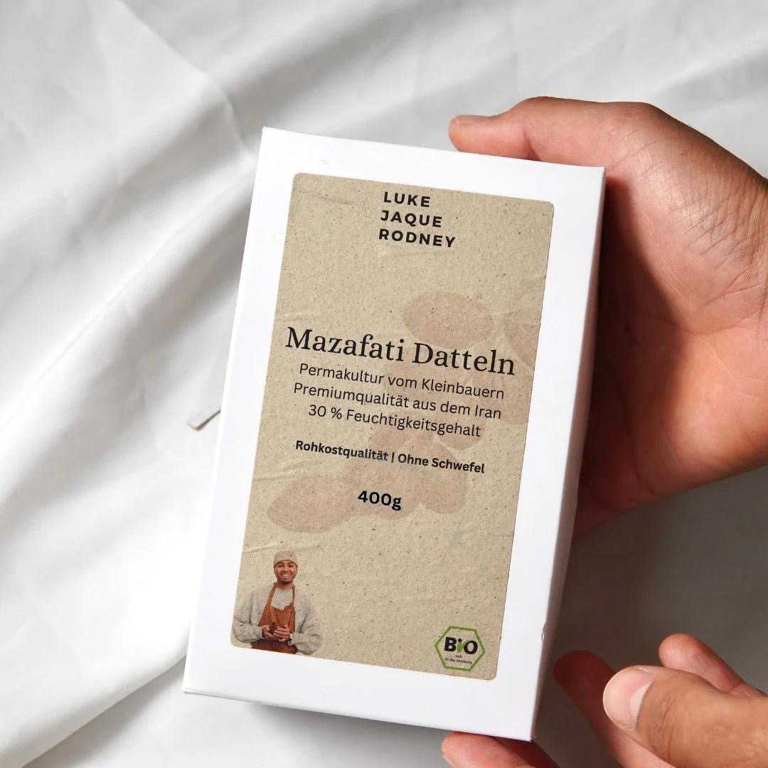 Eine Hand hält eine weiße Schachtel von Datteln | Mazafati Permakultur von Luke Jaque Rodney Store, auf der das deutsche Etikett mit biologischen Mazafati-Datteln aus dem Iran, Zertifizierungsangaben, Produktinformationen und ein kleines Foto einer Frau zu sehen sind.