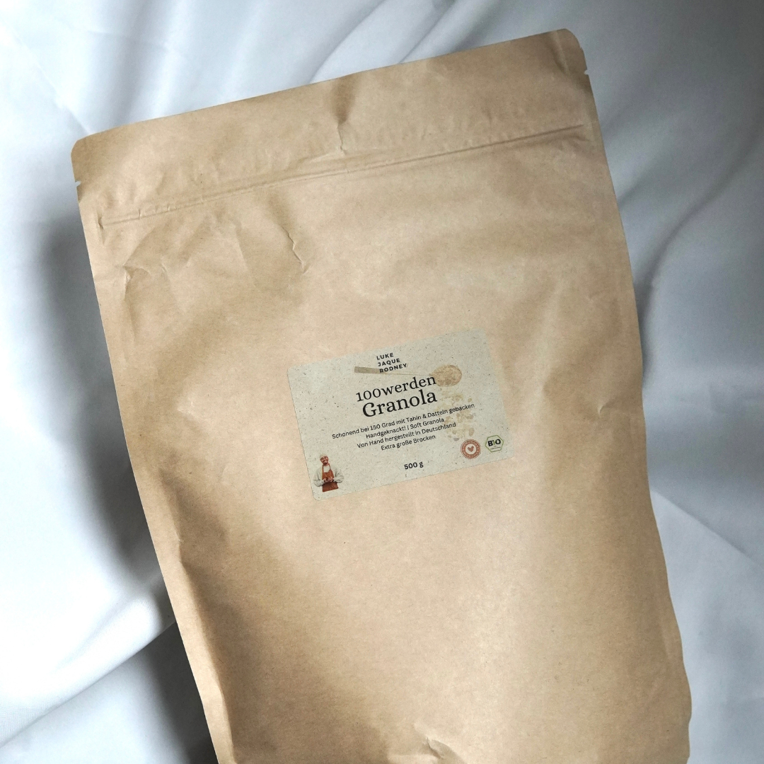 Eine große braune Papiertüte mit der Aufschrift "100 werden Granola" aus dem Luke Jaque Rodney Store, auf der Ceylon Zimt und eine Produktinformation mit einer kleinen Flaschenabbildung abgebildet sind, liegt auf einer weißen Fläche.