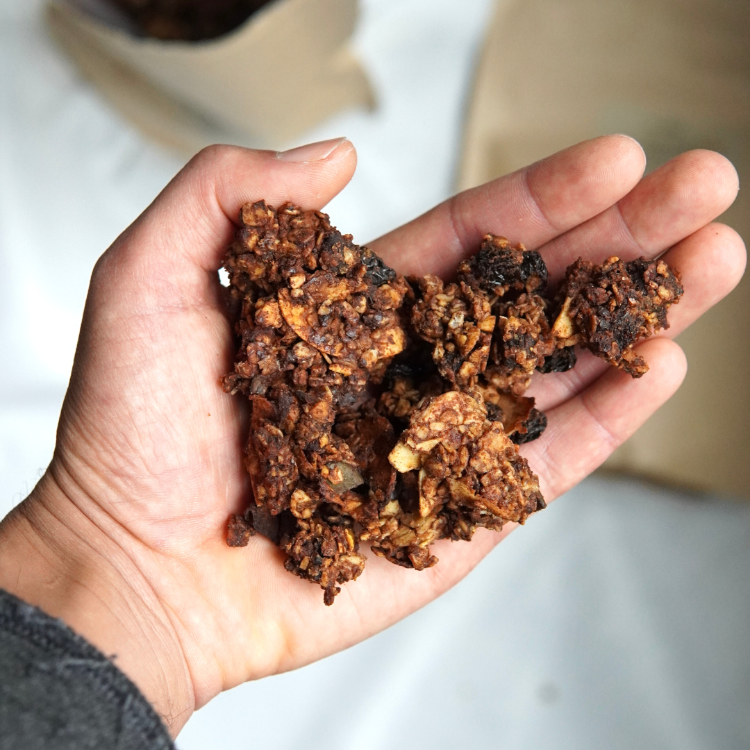 Eine Hand hält Cluster von dunklem, knusprigem 100 werden Granola mit subtilen Tahin-Noten aus dem Luke Jaque Rodney Store vor einem verschwommenen Hintergrund.