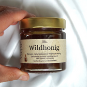 Eine Hand hält ein Glas mit Wildhonig | Deutscher Rohhonig aus dem Luke Jaque Rodney Store, das mit einem goldenen Deckel versehen ist. Auf dem Etikett steht, dass dieser Premiumhonig rein, natürlich, mit wenig HMF und reich an Enzymen und Mineralien ist.
