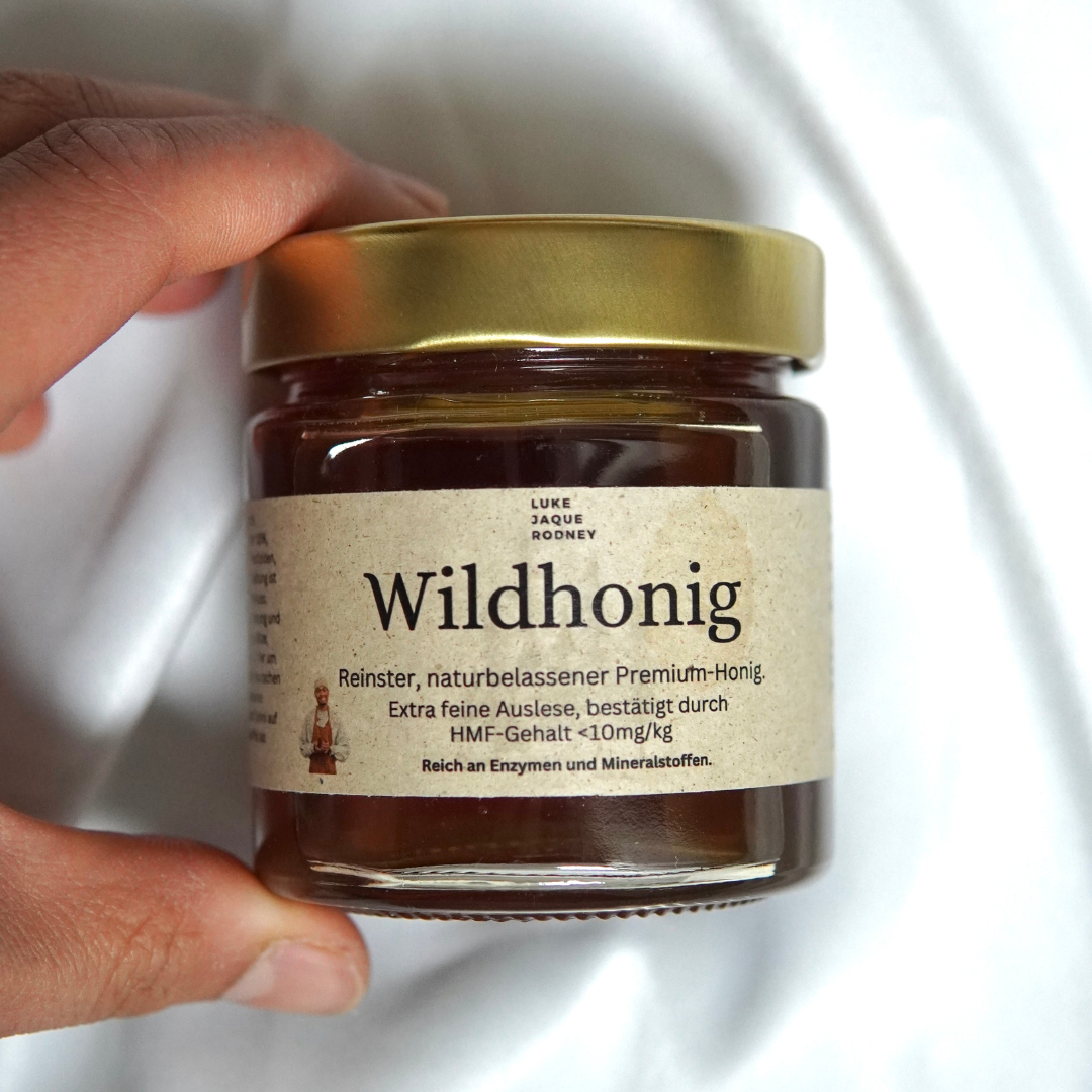 Eine Hand hält ein Glas mit Wildhonig | Deutscher Rohhonig aus dem Luke Jaque Rodney Store, das mit einem goldenen Deckel versehen ist. Auf dem Etikett steht, dass dieser Premiumhonig rein, natürlich, mit wenig HMF und reich an Enzymen und Mineralien ist.