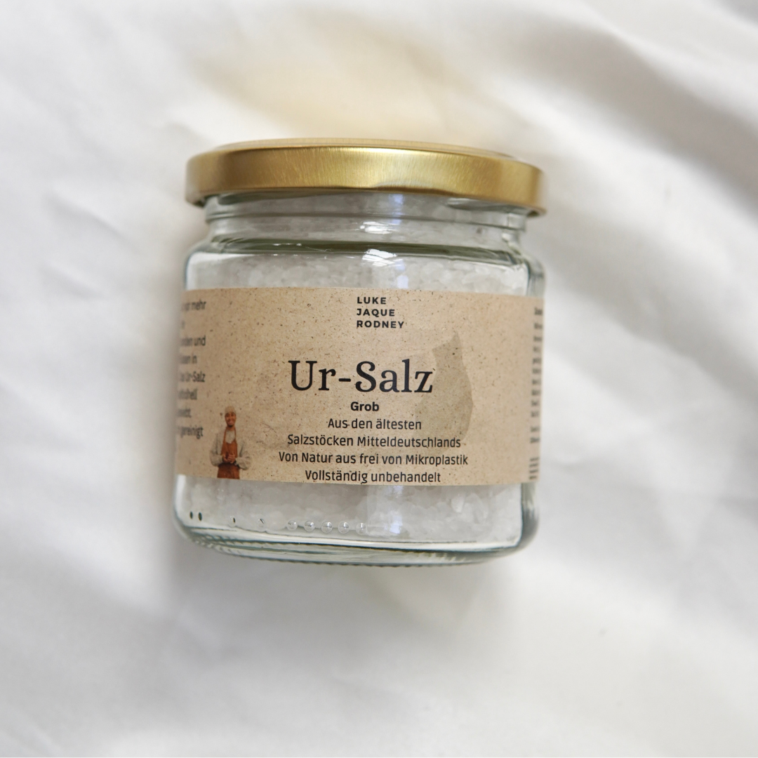 Das Ur-Salz | Frei von Mikroplastik von Luke Jaque Rodney Store wird in einem Glasgefäß mit goldenem Deckel geliefert. Das deutsche Etikett hebt das Salz als rein, unbehandelt und frei von Mikroplastik hervor und unterstreicht damit seine natürliche Herkunft.