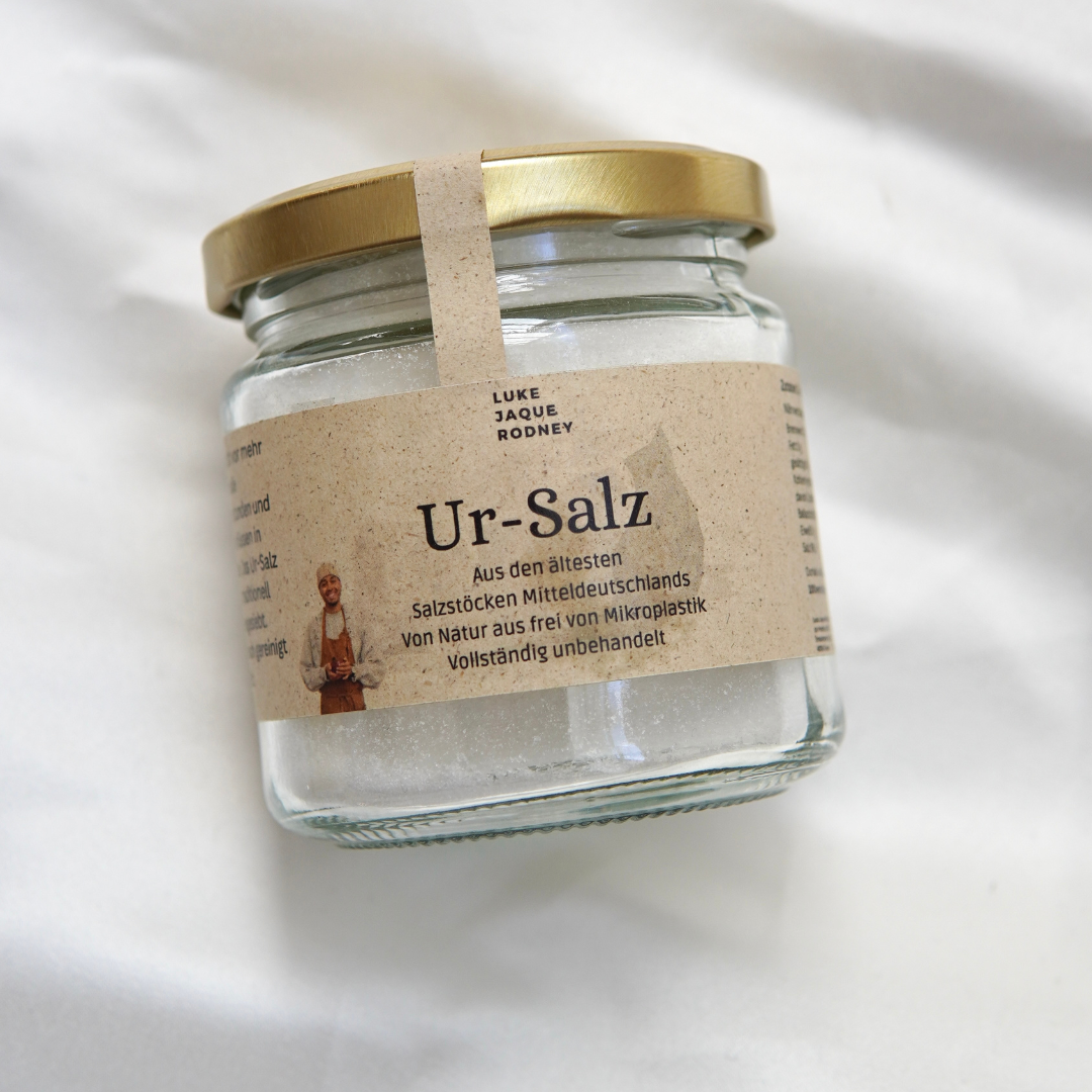 Ein Glasgefäß mit goldenem Deckel aus dem Luke Jaque Rodney Store enthält Ur-Salz | Frei von Mikroplastik, mit einem deutschen Etikett, das das Salz als natürlich rein, unverarbeitet und frei von Mikroplastik beschreibt.