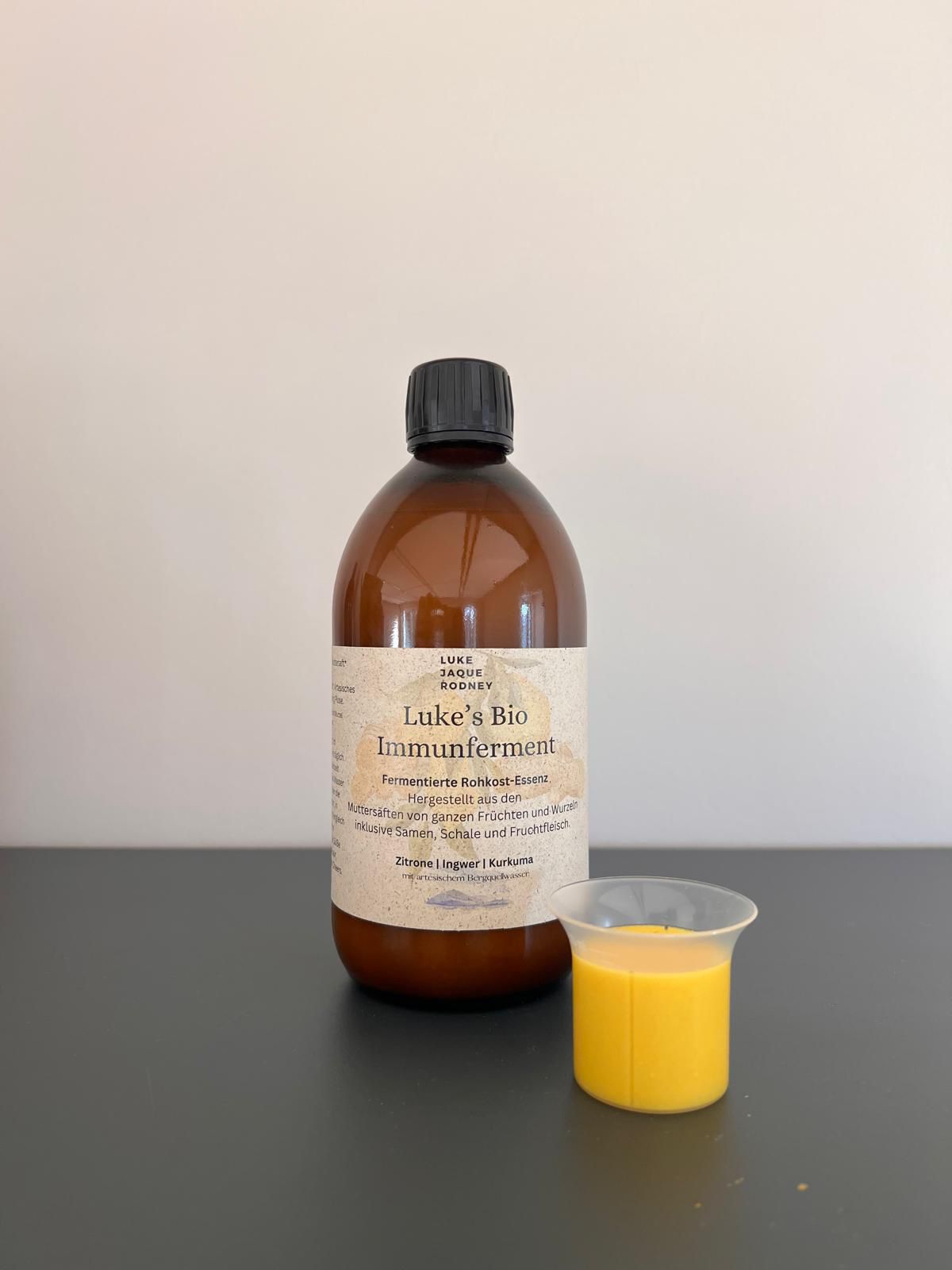 Eine braune Glasflasche mit der Aufschrift "Lukes Bio Immunferment I 100% RAW" aus dem Luke Jaque-Rodney Store steht auf einer dunklen Fläche neben einem kleinen Plastikbecher mit gelber Flüssigkeit vor einem schlichten, hellen Hintergrund.