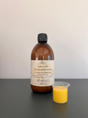 Eine braune Glasflasche mit der Aufschrift "Lukes Bio Immunferment I 100% RAW" aus dem Luke Jaque-Rodney Store steht auf einer dunklen Fläche neben einem kleinen Plastikbecher mit gelber Flüssigkeit vor einem schlichten, hellen Hintergrund.