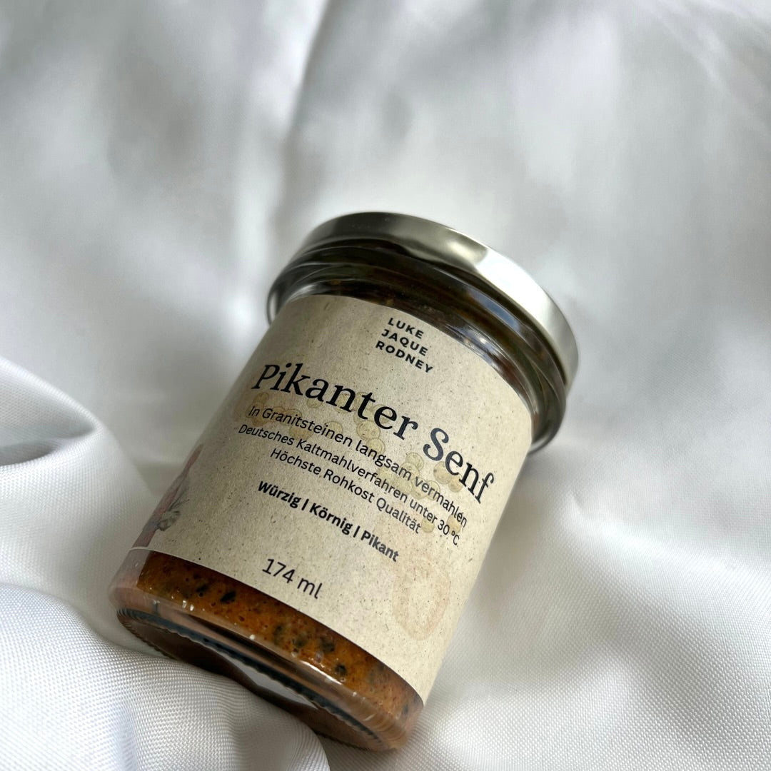 Ein Glasgefäß von Echter Senf | Rohkost von Luke Jaque Rodney Store mit silbernem Deckel und beigem Etikett mit deutschem Text steht auf weißem Stoff. Der 174 ml große Senf zeichnet sich durch seine Rohkost-Qualität aus.
