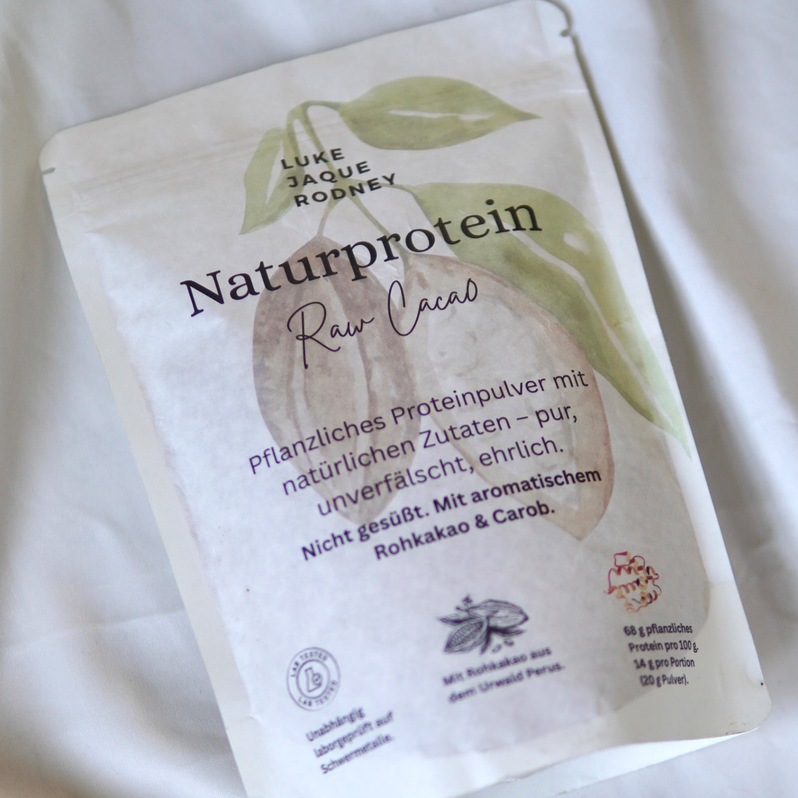 Eine Packung Luke Jaque-Rodney Naturprotein (Raw Cacao) Proteinpulver mit deutschem Text, Bildern von Blättern und Kakao und Zertifizierungen auf weißem Hintergrund - ein veganes, rohes Produkt mit Erbsenprotein.