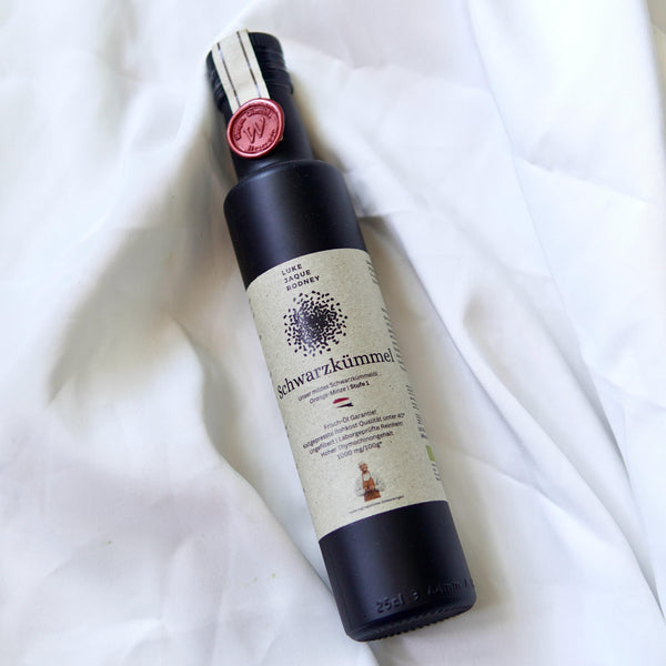 Eine dunkle Flasche mit der Aufschrift "Schwarzkümmelöl" von Luke Jaque-Rodney ruht auf weißem Stoff. Mit rotem Siegel und minimalistischem Design bietet sie rohe Qualität und natürlich hohen Thymochinongehalt.
