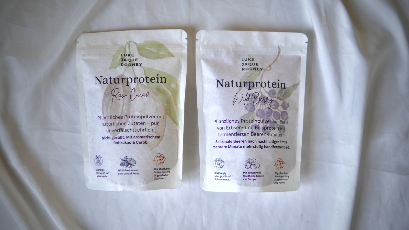Zwei weiße Naturprotein-Beutel von Luke Jaque-Rodney liegen auf einer weißen Fläche: einer mit der Aufschrift Wild Berry und einer mit einer lila Beerengrafik, der andere mit einem grünen Kakao-Motiv. Beide tragen einen deutschen Text.